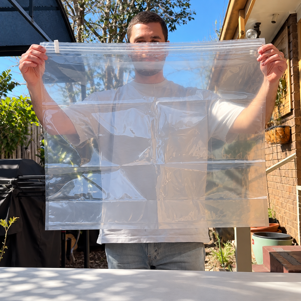 A man outdoors holds up a large clear Brining and marinating bag zippered storage bag with multiple horizontal and vertical compartments. The background shows a residential backyard with a brick wall, plants, and a barbecue grill.